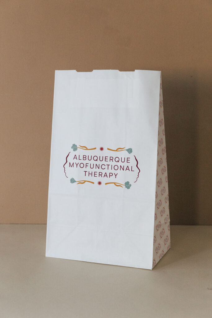 Branded paper bag featuring the Albuquerque Myofunctional Therapy submark printed on the front.